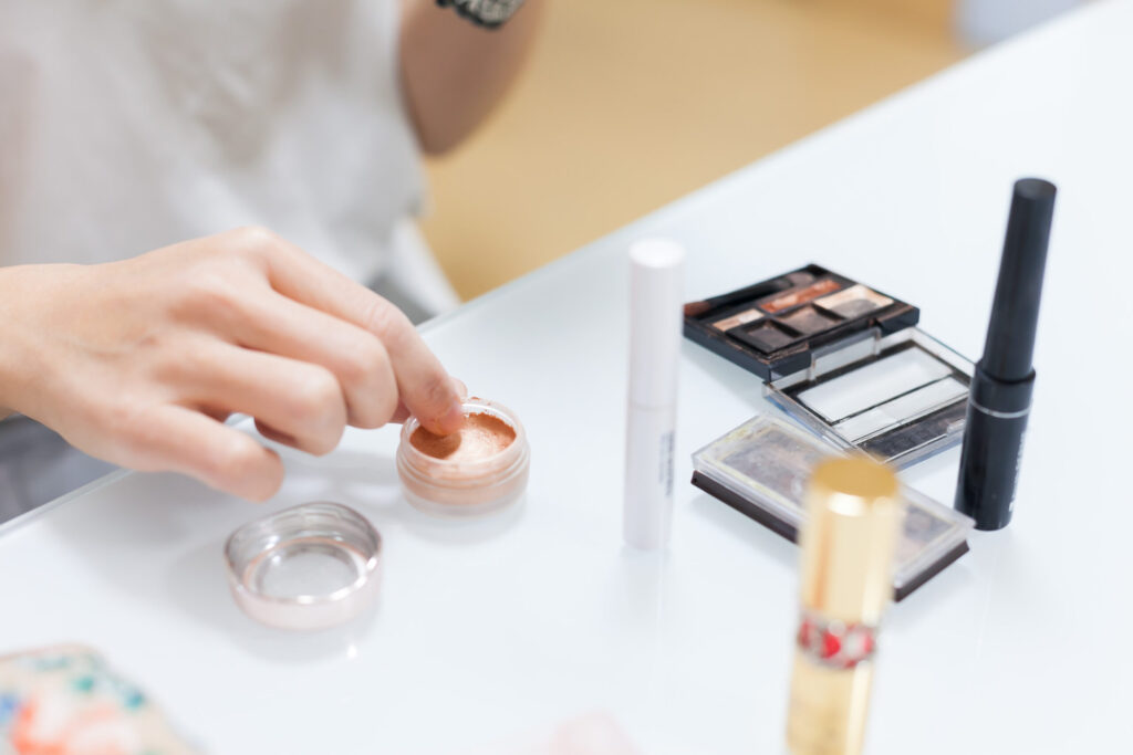 アイシャドウ、口紅などの化粧品が、真っ白なテーブルに置かれている。アイシャドウを指に取っている女性の手元が見える。