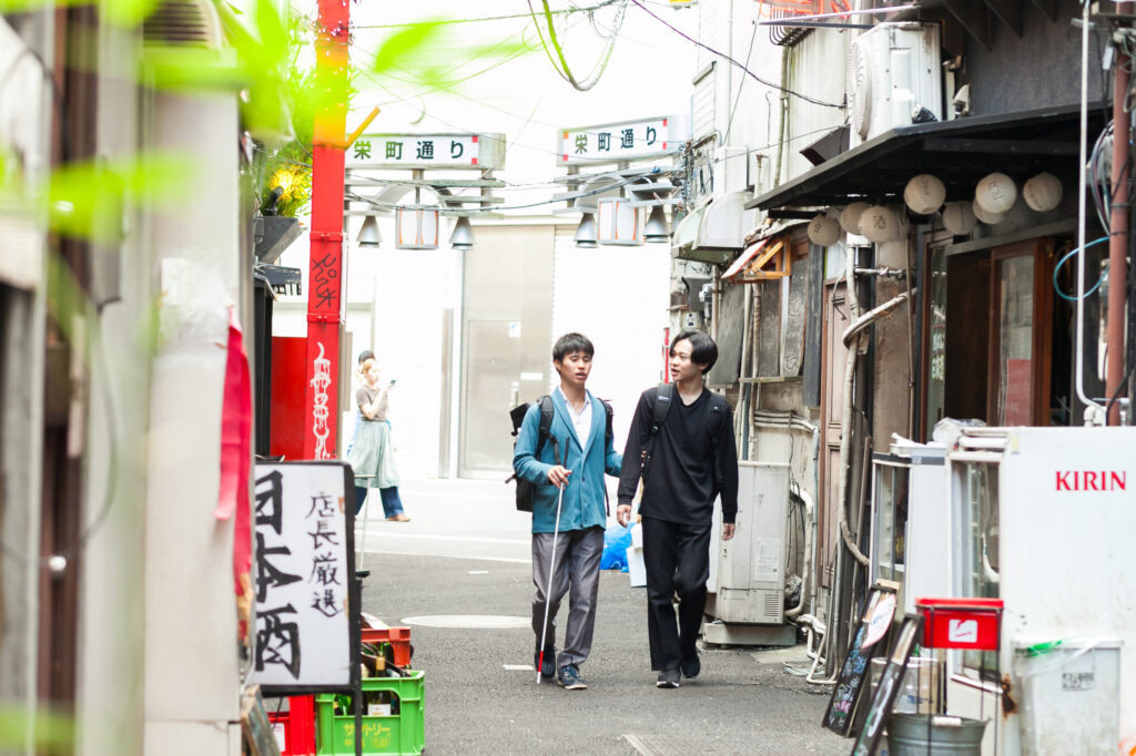 白杖の男性とガイドヘルパーが、飲食店が並ぶ都市部の古い裏路地を会話をしながら歩いている。