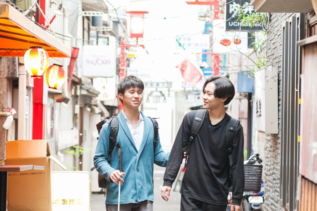 白杖の男性とガイドヘルパーが、日中に都会の裏路地のような飲食店街を歩いているところ。