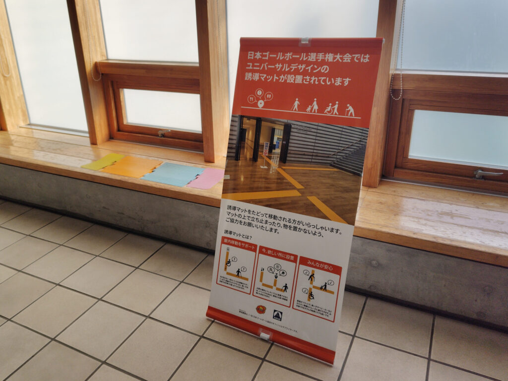 簡単に設置できる「誘導マット」の見本と紹介パネル。パネルには「日本ゴールボール選手権大会ではユニバーサルデザインの誘導マットが設置されています」と書かれていて、誘導マットの解説イラストや、誘導マット上で立ち止まったり物を置いたりしないように注意喚起がされている。
