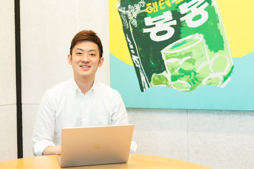 高橋さんが、オフィス内の打合せスペースで、こちらを見て座っている。デスクには開かれたノートパソコンがある。