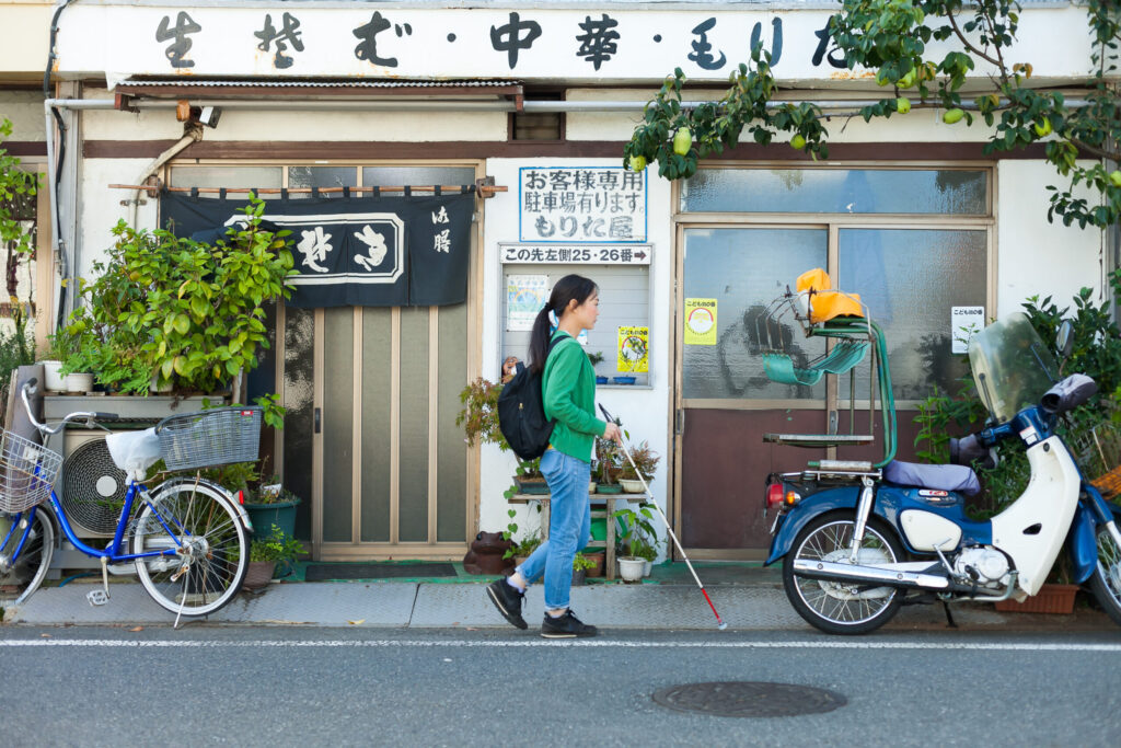 住宅街を白杖を持って歩いている女性。歩道がなく自転車やバイクが止められている。背景には「生そば、中華」と書かれた看板の店。