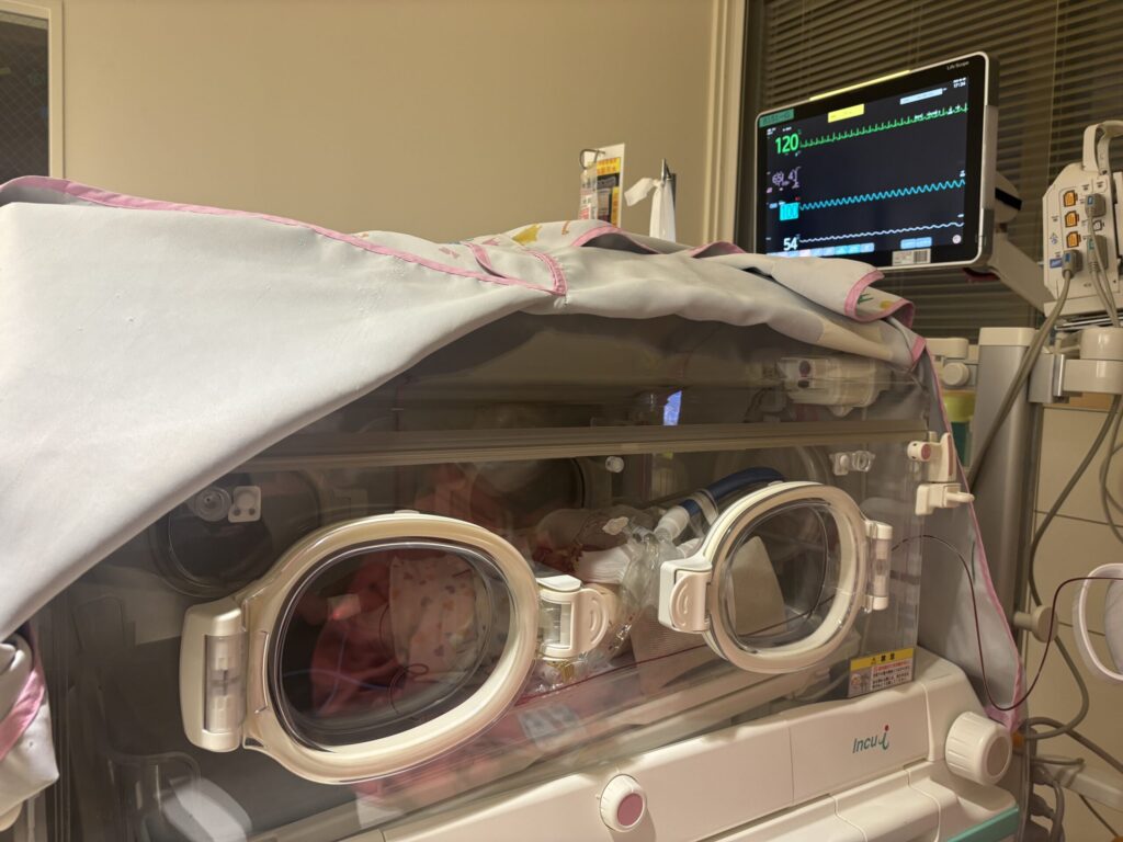 NICUの保育器を外から見た様子。心電図などを写すモニターがあり、透明な箱状の保育器には、保護者や医療者が手を入れられる楕円状の扉がついている。保温のためか、布がかぶせられている。