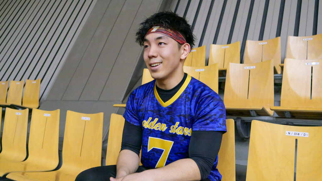 観客席に座って、笑顔でインタビューに答える金子選手。青地に黄色いアクセントのユニフォームを着ている。