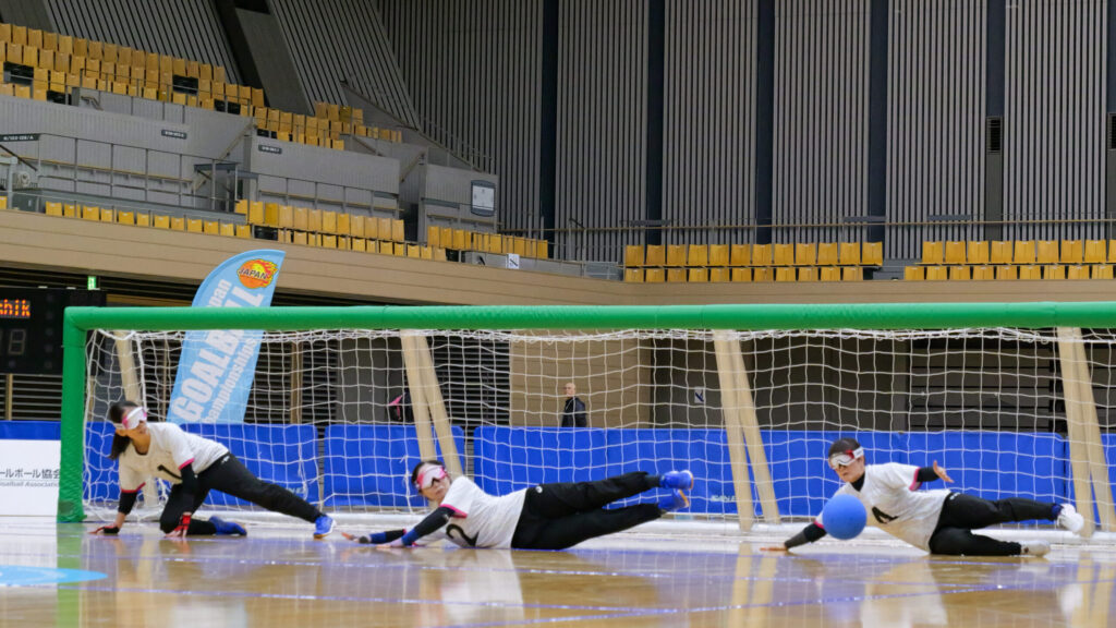 3人の選手がゴール前でディフェンスをしているところ。全員が右向きに低く飛び、寝そべるような姿勢で並んで、ゴール全体をガードしている。