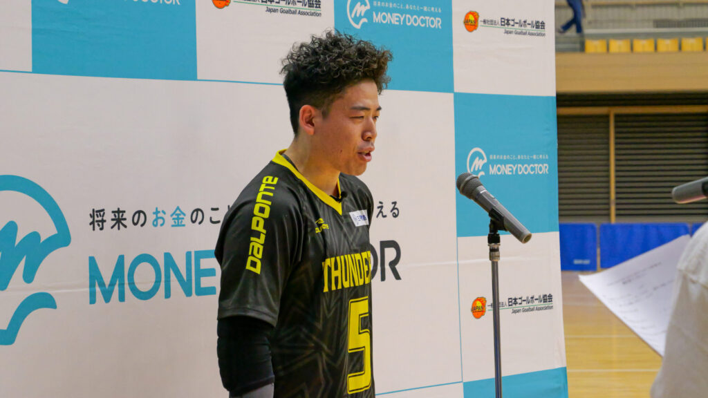 大会パネルの前で、マイクに向かってインタビューに答えている山口選手。黒地に鮮やかな黄色のアクセントが入ったユニフォーム。