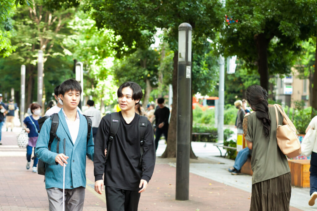 人通りの多い都会の広い歩道をヘルパーと白杖の男性が歩いている。