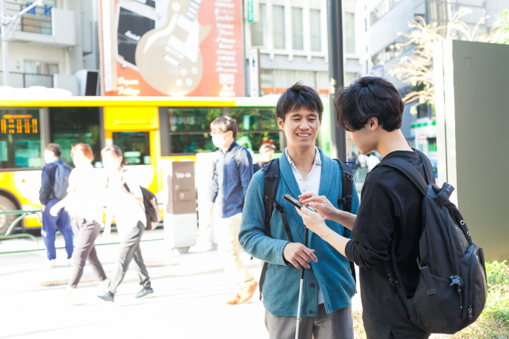 白杖の男性とヘルパーの男性が都心の歩道の隅で立ち止まって話している。ヘルパーはスマートフォンで何かを調べている。