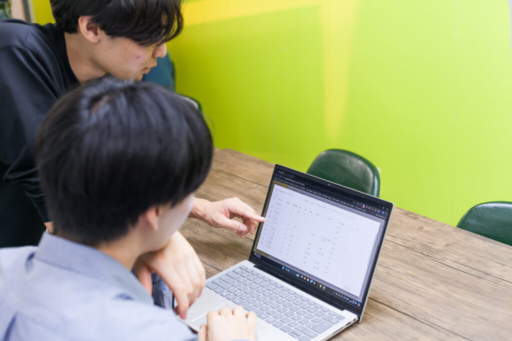 座ってノートパソコンを見ている男性と、その左隣から、立って画面を指さし話しかけている男性。