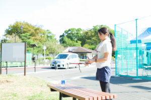 スポーツウエアを着た視覚障害者の女性が、運動場のそばにあるベンチの前に立ち、折り畳み式の白杖を畳んでいるところ。