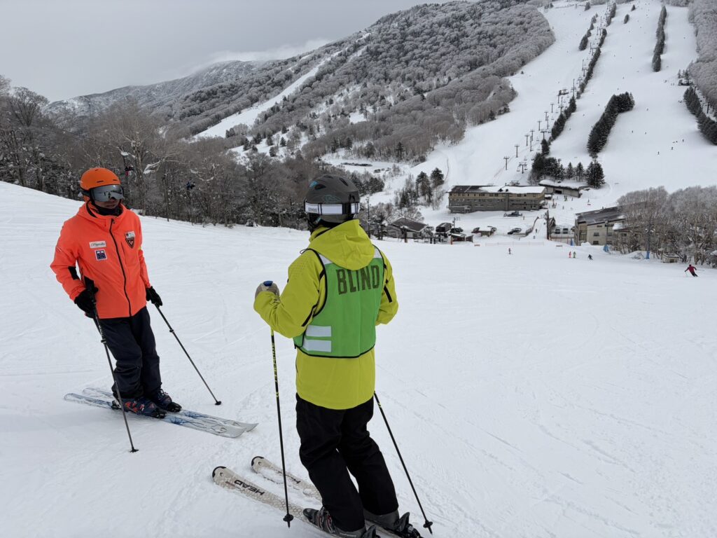 ケビンさんとインストラクターが、斜面に止まって会話をしているところ。かなり急な斜度のコースで、下の方にリフト乗り場が見える。