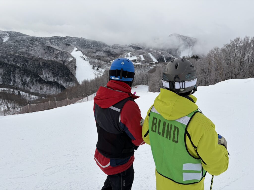 ガイドさんと筆者ケビンさんが、コース斜面の上の方から、これから滑る斜面を確認している。コースの奥には、雪の山々が見える。