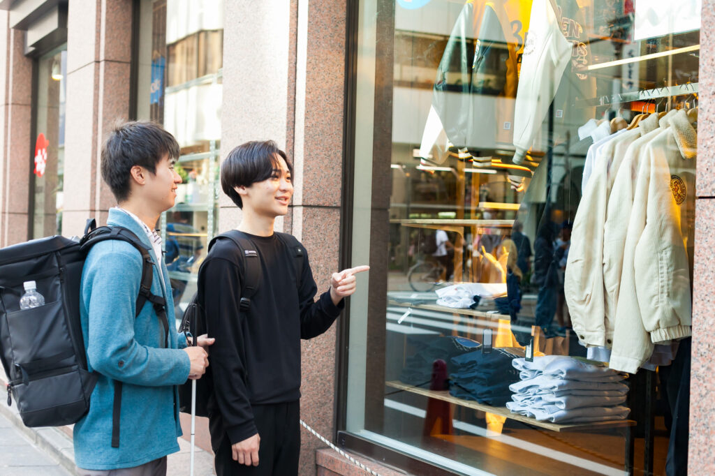 視覚障害者とガイドヘルパーが、洋服店のウィンドウを外から眺めている。ヘルパーは指をさしながら商品の説明をしている。