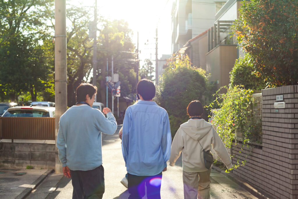 男性二人と女性一人が住宅街を並んで歩いている後姿、夕日が沈む方向に向かっ話しながら歩いている。