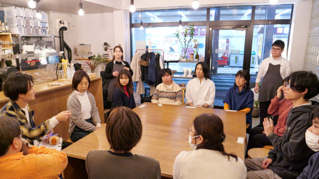 大きな木製のテーブルを囲んで座る参加者と主催の設楽さん。全員話している設楽さんの方を向いている。カフェスタッフ2人が参加者の輪の外側に立っている。