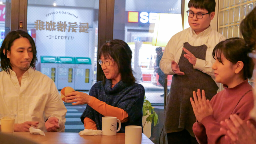 話し終わった白いシャツの男性参加者から次の青いニットを着た女性の参加者へボールが渡されているところ。周囲の人は拍手をしている。