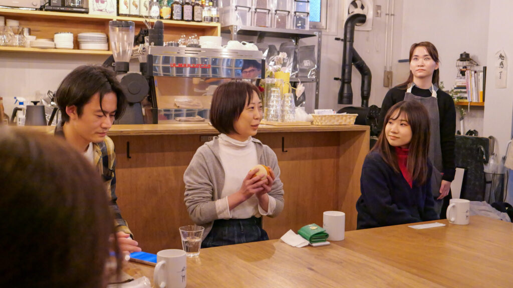 白いタートルネックにグレーのカーディガンを羽織った女性が、両手に布製のボールをそっと持ちながら話している。周囲の人はリラックスした様子で耳を傾けている。