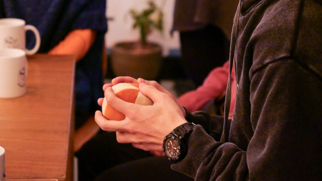 布製のボールを両手で包むように握っている黒っぽい服を着た男性の手元のクローズアップ。木製のテーブル、周囲の人の手元も見える。