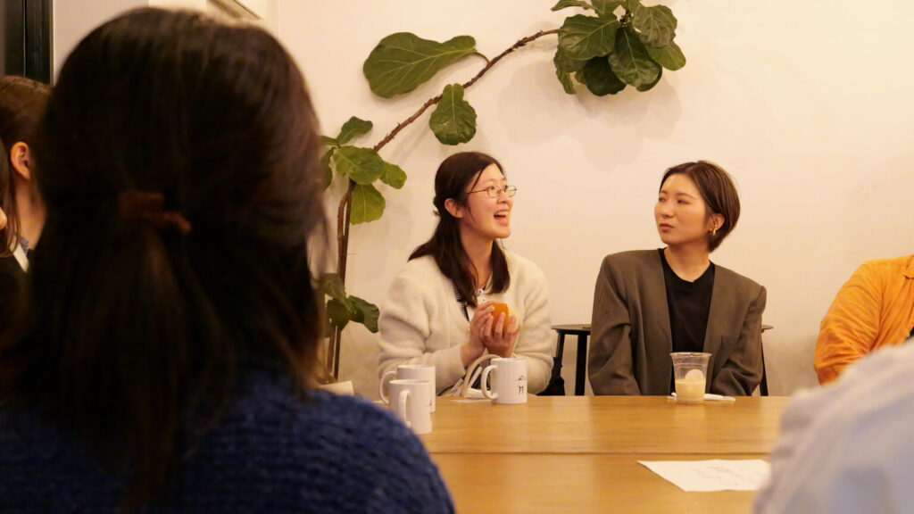 布のボールを持って、笑顔で楽しそうに話している白いセーター姿の女性。背景にはカフェの観葉植物の大きな枝と葉が見える。