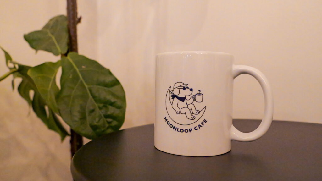 丸くて黒っぽいテーブルに置かれた、白いマグカップ。カップには「MOONLOOP CAFE」のロゴと、三日月に腰かけてマグカップを持った犬のイラストが描かれている。