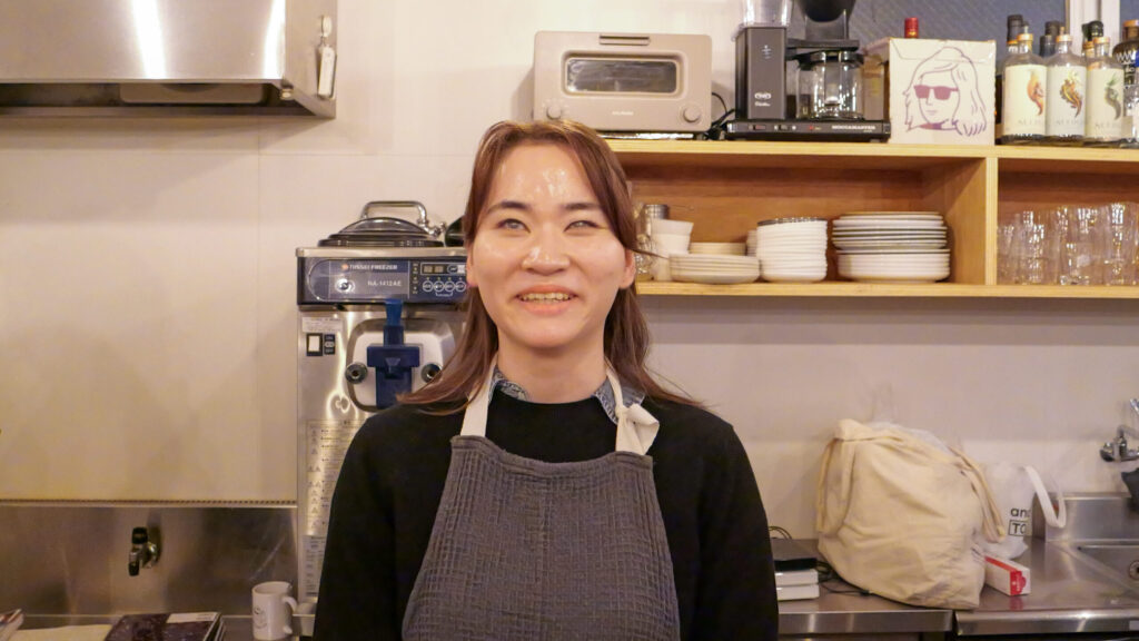 キッチンを背景にこちらを向いて笑顔で話している隅本さん。黒い服にグレーのエプロン姿。