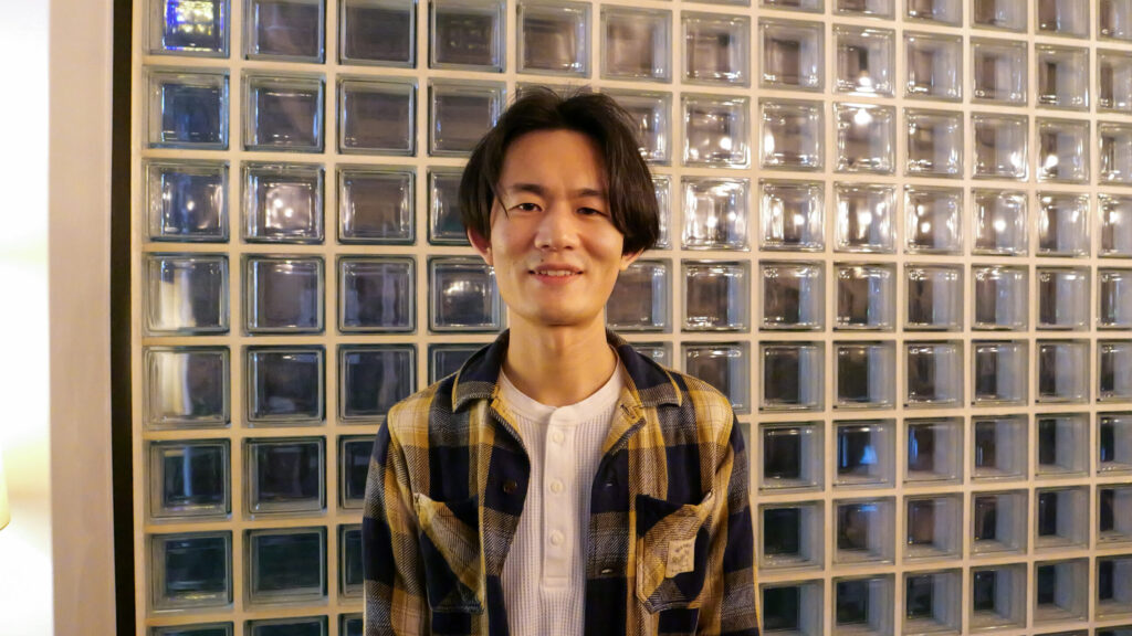 ブロックガラスを背景に、こちらを向いて笑顔で話している設楽さん。白いTシャツに黄色と黒のチェック柄のシャツを羽織っている。