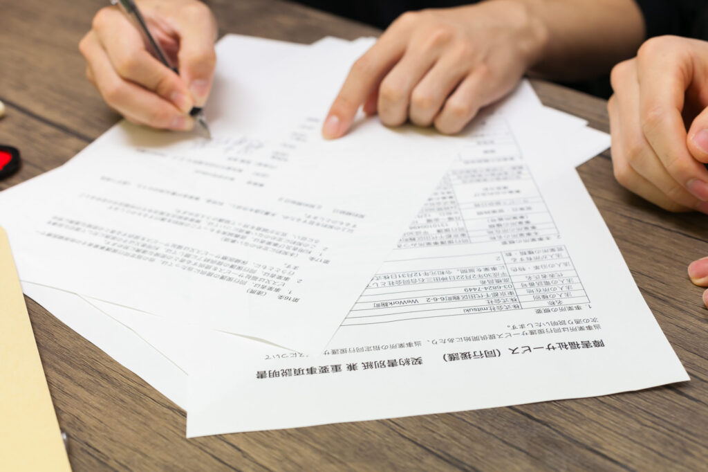 机の上の複数の書類と人の手元の写真。左手で指さし確認をしながら右手にペンを持ち書類に書き込んでいる様子。