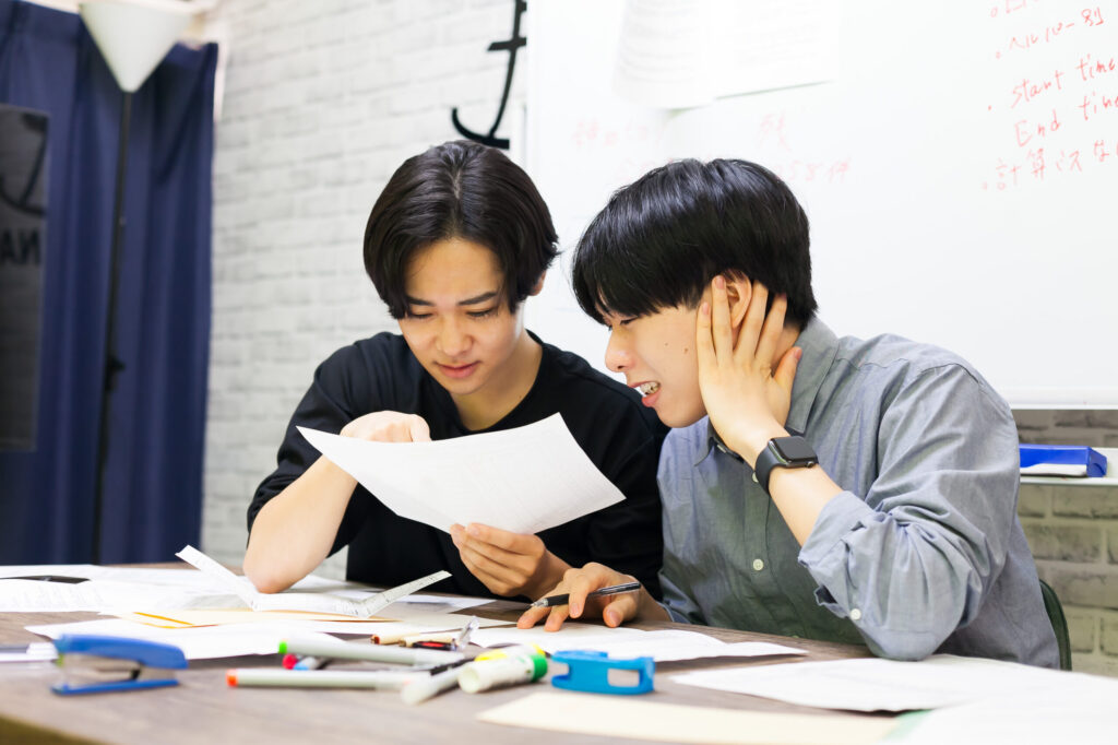 男性二人が机の上に書類やペンなどを広げて話している。一枚の書類を二人でのぞき込み、確認しながら困った顔をしている。