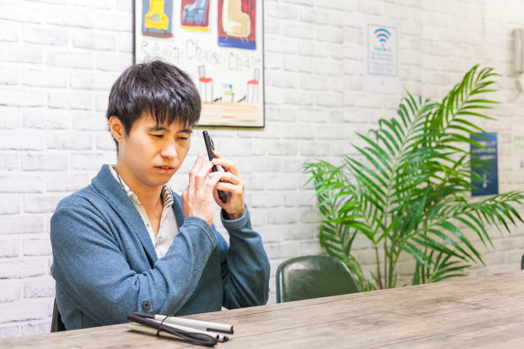 視覚障害者がテーブルを前に椅子に座っている。スマートフォンを操作しながら耳元に持ち、音声読み上げで読んでいる。