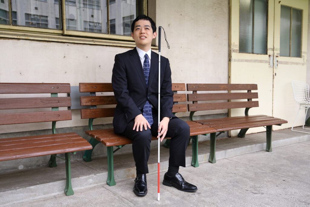 紺色のスーツ姿で木製のベンチに腰かけている濱田さん。足の間に白杖を立てかけて左手を添えている。