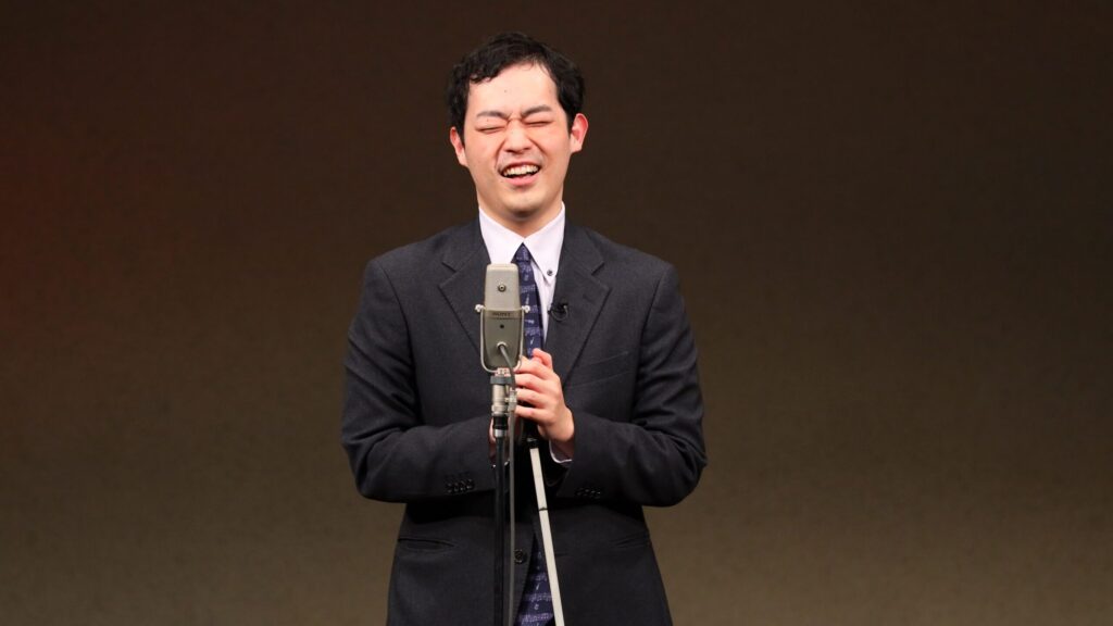 舞台で公演中の濱田さん。クシャッとした笑顔でスーツ姿。マイクの前に立ち、白杖を両手で包むように持っている。