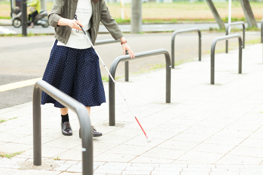 白状を持った女性が、公園などの入り口にある車両侵入防止の柵を、手で確認しながら慎重に歩いている様子。