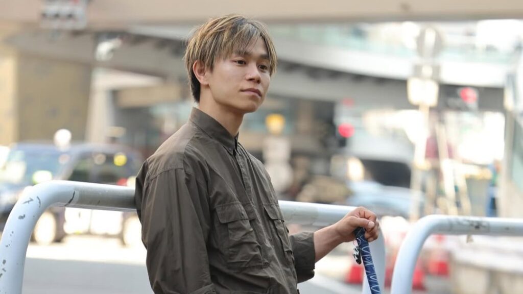 都心部の歩道で、茶色っぽいシャツを着て白杖を持って立っている濵ノ上選手。