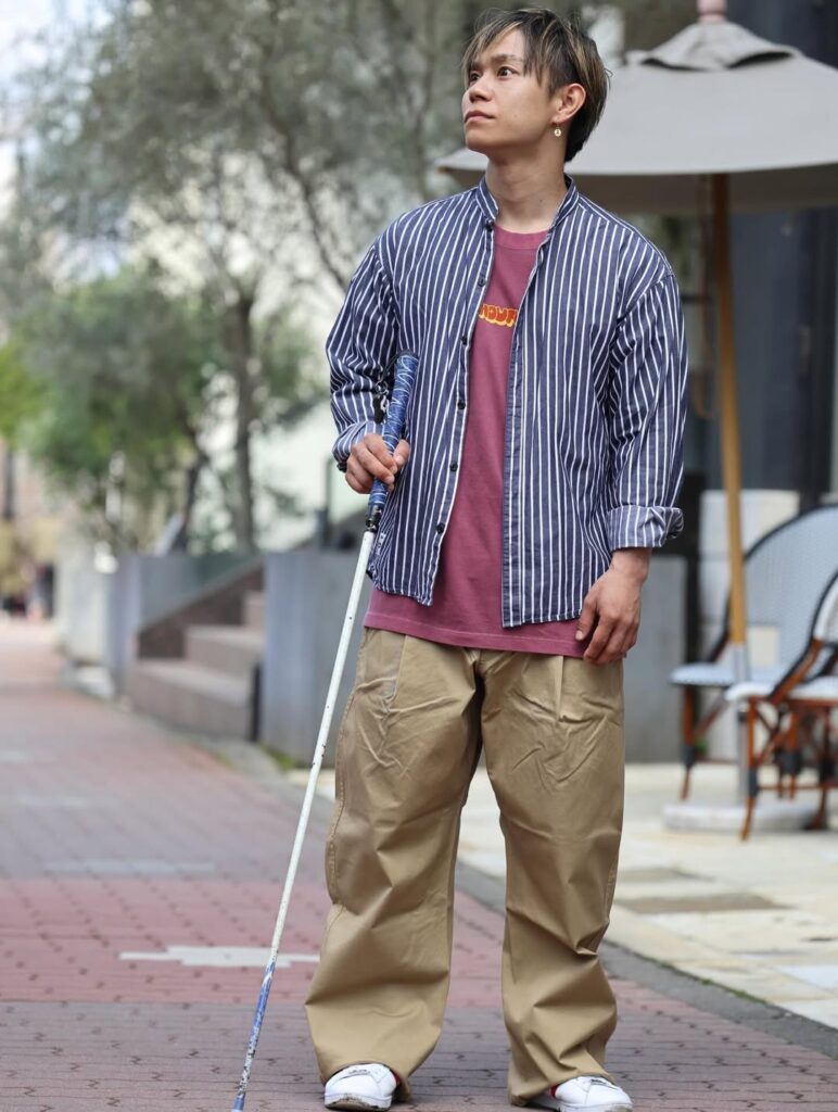 街でカジュアルな服装で白杖を持って立っている濵ノ上選手。紫系のTシャツの上に紺地に白の縦ストライプのシャツを羽織っている。ボトムスはベージュ系のチノパン風。靴は白いスニーカー。左耳にピアスが光っている。