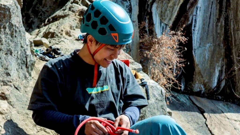 岩場で赤いクライミングロープを準備している濵ノ上選手。明るい青のヘルメットをかぶり、紺色のTシャツとブルーのボトムス姿。