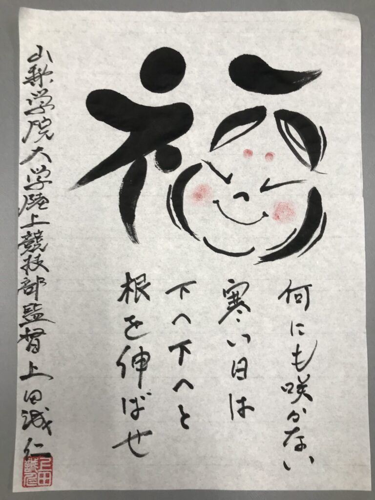 半紙に筆で書かれた文字。お多福の顔がデザインされた「福」の漢字。その下に「何にも咲かない寒い日は、下へ下へと根を伸ばせ」言葉。半紙の左側に「山梨学院大学陸上競技部監督上田誠仁」の文字。
