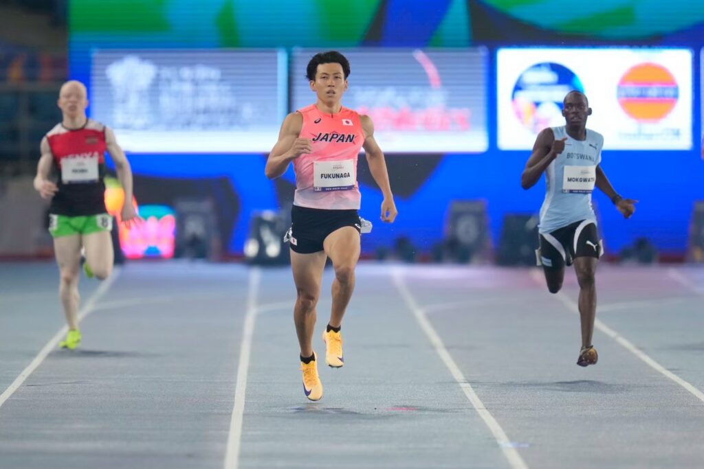 トラックを走っている競技中の福永選手。赤いトップスと黒系の短パンのユニフォーム姿。両隣には海外の選手の姿も見える。