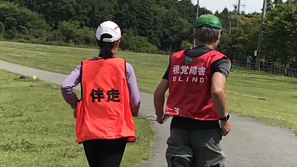 「視覚障害」のビブスを付けたランナーと、「伴走」のビブスを付けたガイドランナーが公園内を走る様子。
