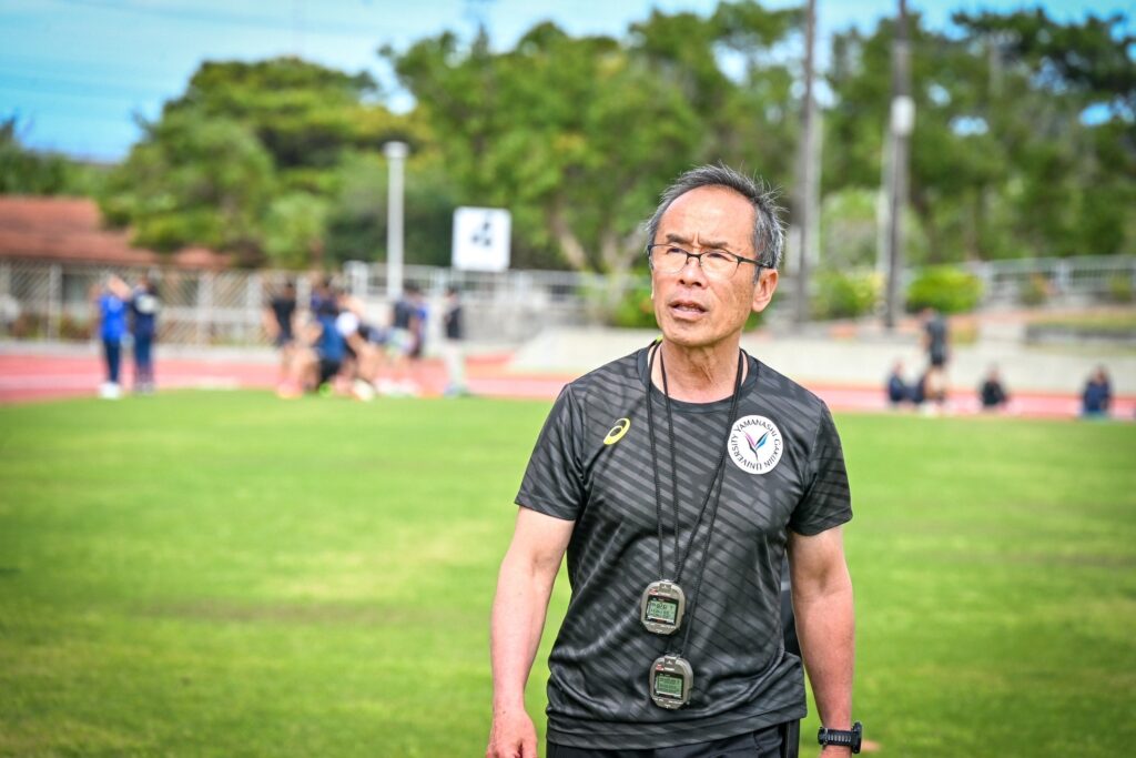 芝生の競技場で、黒いスポーツウエア姿、ストップウォッチをふたつ首から下げて、眩しそうに右上を見ている上田監督。