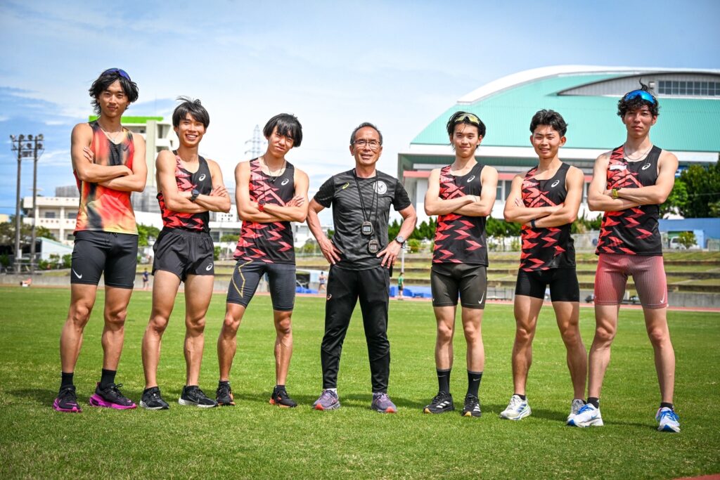 芝生の競技場で、腰に手を当てた上田監督の左右に三人ずつ男性の陸上競技の教え子が立っている。