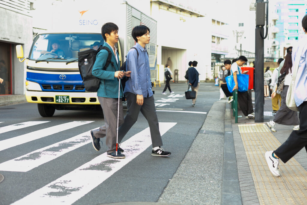 白杖の男性とガイドヘルパーが横断歩道を渡り終えるところ。車道にはトラックが停止している。