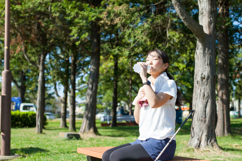 女性が公園のベンチでペットボトルの水を飲んでいる画像。脇に白杖が置かれている。