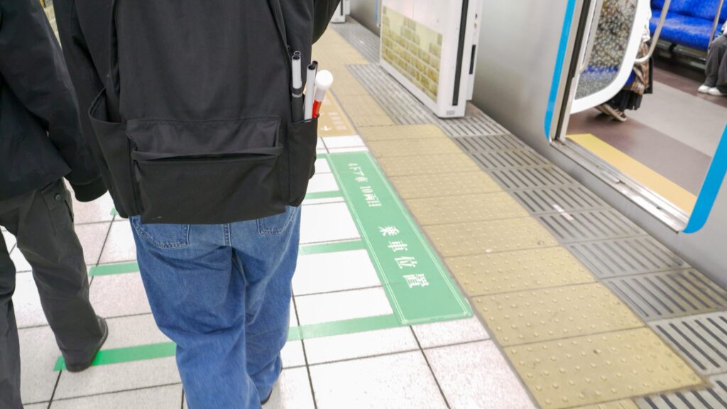 駅のホームを歩く秋山さんと木村さんの足元を後ろから写した写真。秋山さんのリュックのサイドポケットに折り畳み式の白杖が入っている。ホーム端には点字ブロックがある。