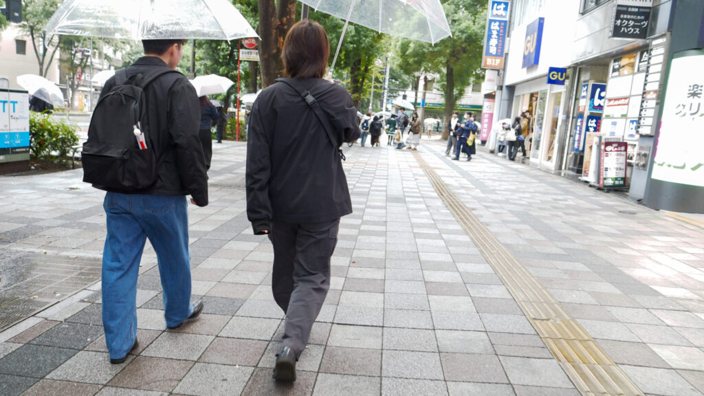 傘をさして、繁華街の広い歩道を並んで歩く木村さんと秋山さんの後姿。