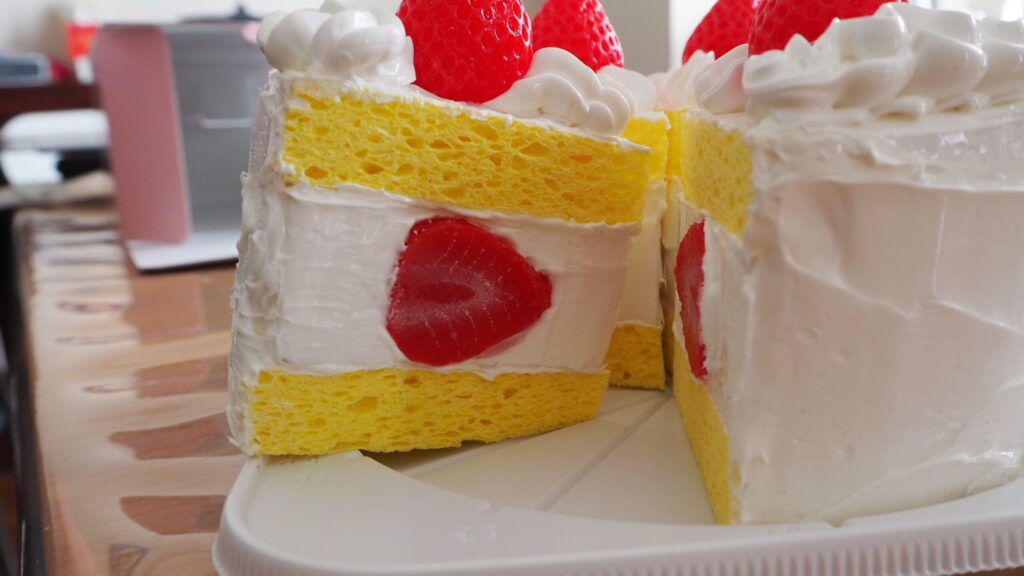 いちごのショートケーキの食品サンプル。白い生クリームのデコレーションが全体にほどこされ、赤いイチゴが乗っている。5ピースあり、並べるとホールケーキの一部が欠けた形になる。各ピースの断面は黄色いスポンジケーキが見えていて、スポンジとスポンジの間にクリームとイチゴが挟まっている。