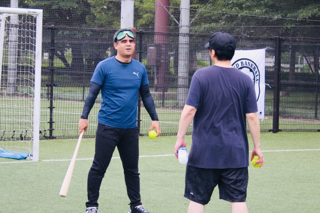 右手にバット左手にボールをもった視覚障害者の男性が、向き合って立つ男性と話している。