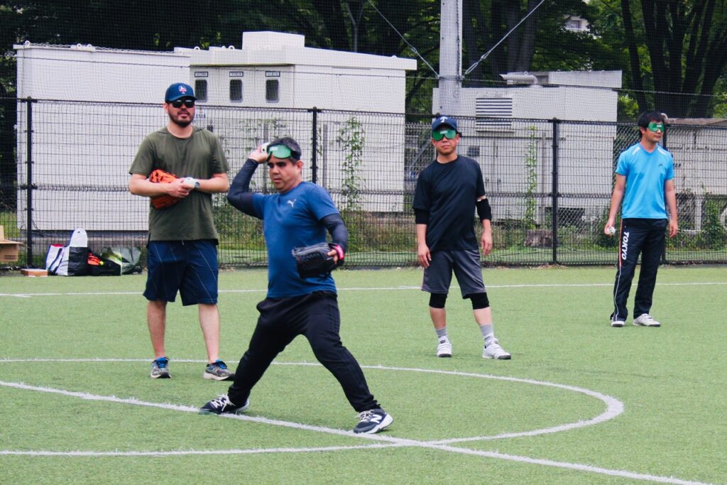 視覚障害者の男性が、コーチに投球フォームの指導を受けている様子。周囲にはアイマスクをした、ほかのプレーヤーが2人立っている。