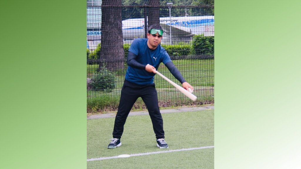 男性が、右手にバット左手にボールを持ち、右手の肘を曲げ左手はピンと伸ばしている。バットの先にボールを当てながらバッティングの準備に入っている。