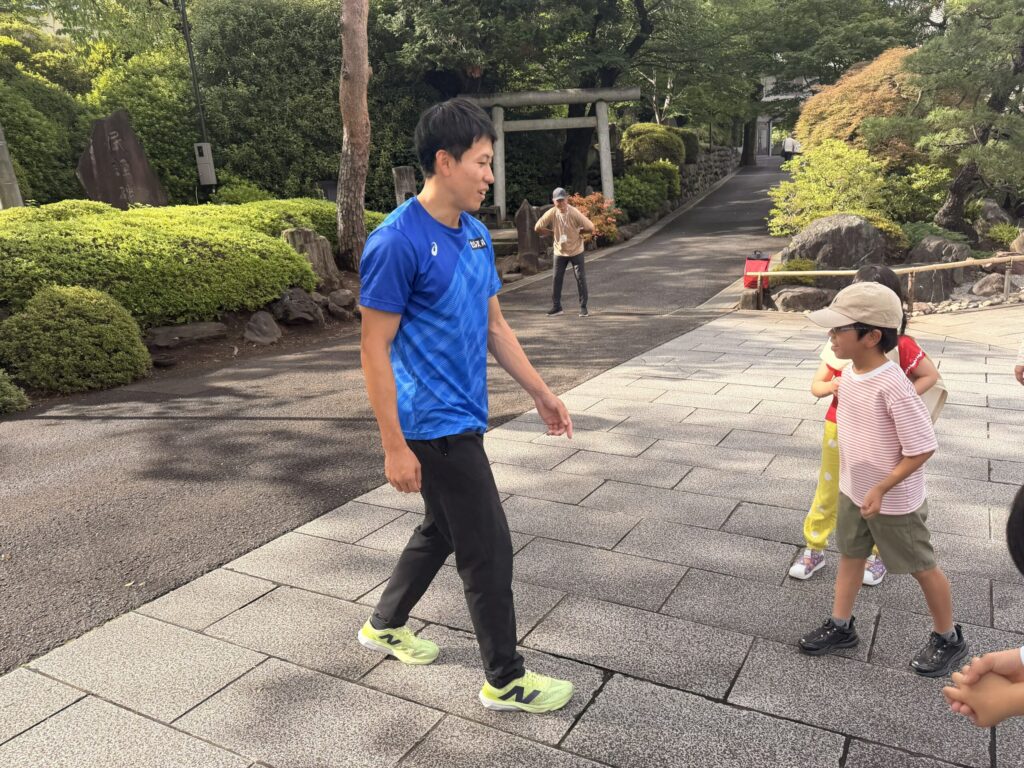 青いスポーツウエアを着た福永選手が屋外で子どもに指導している様子。片足を踏み出していて子どもたちがまねしている。