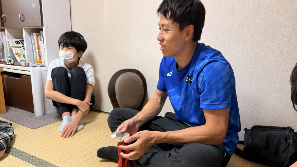 畳の上で胡坐で座り、銀メダルを手に中学生と話している福永選手。